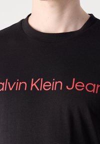 Černé bavlněné tričko s kulatým výstřihem. Na přední straně zdobeno výrazným červeným nápisem "Calvin Klein Jeans". Hladká struktura.