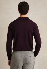 Man met donker krullend haar draagt een donker bordeauxrode blouse met lange mouwen en grijze gestructureerde broek, staat met één hand in de zak, met de rug naar de kijker gekeerd.