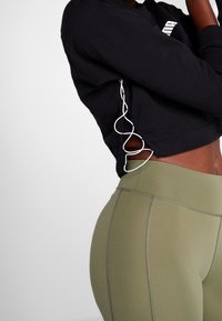 Felpa nera corta con dettagli in nastro sui lati, abbinata a leggings verde oliva a vita alta con accenti cuciti.