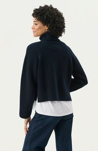 Navy ribbed sweater with a high collar and cropped design, featuring long sleeves and a textured knit pattern. Worn over a white shirt.