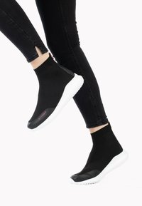 Czarne buty typu slip-on z plecionym wierzchem i białą gumową podeszwą; wyróżniają się kontrastującą, czarną osłoną palców oraz czerwonym detalem z logo.