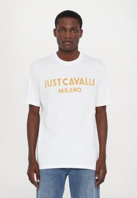 Bílá bavlněná tričko s krátkými rukávy, na přední straně s výrazným žlutým nápisem "JUST CAVALLI MILANO". Jednoduchý, volný střih.