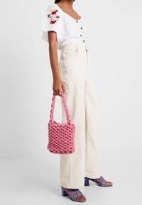 Bolso de tela tejido en rosa con un asa trenzada, combinado con una blusa blanca de botones y pantalones de canalé de cintura alta.