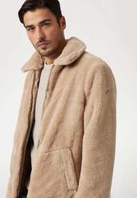 Giacca in pile beige con una texture morbida, chiusura con zip e un ampio colletto. Presenta una tasca laterale e un piccolo logo sul manico.