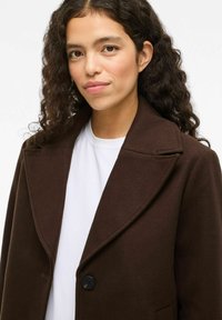 Una mujer con cabello rizado y oscuro lleva un abrigo marrón oscuro sobre una camisa blanca, frente a un fondo claro y liso.