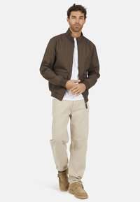 Veste bomber brun avec un col montant, fermeture éclair à l'avant et poches latérales, portée avec un t-shirt blanc et un pantalon beige, associée à des bottes tan.