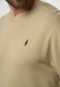 Kreminės spalvos medvilninė marškinė su apvaliu kaklu ir mažo tamsiai mėlyno siuvinėto figūros ant kairiojo krūtinės šono. Minkšta tekstūra.