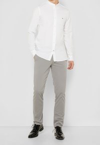 Camicia bianca a maniche lunghe con colletto alla coreana, chiusura con bottoni e piccolo logo ricamato, abbinata a pantaloni grigi chiari e scarpe nere.