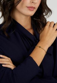 Bracciale d'oro con charm a forma di cuore, design a perline, indossato su un polso. Blusa blu scuro visibile sullo sfondo.