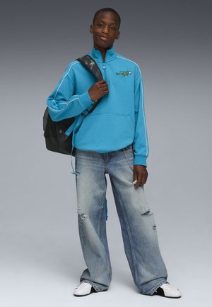 Joven con sudadera azul de cremallera hasta el pecho y vaqueros holgados desgastados, sosteniendo la correa de una mochila negra, de pie frente a un fondo gris.