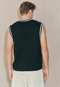 Schwarze, gestrickte ärmellose Sweater mit weißem Besatz an den Schultern, mit lässigem Schnitt und glatter Textur, getragen über hellfarbigen Hosen.