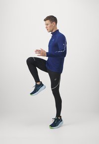 Marineblauw sportshirt met lange mouwen en een halve rits, gecombineerd met zwarte leggings met witte accenten, en blauwe hardloopschoenen met groene zolen.