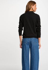 Maglione nero in maglia con maniche lunghe, abbinato a jeans in denim blu a gamba larga con tasche posteriori e cuciture ben visibili.