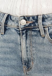 Κοντινό πλάνο ξεθωριασμένου μπλε τζιν με ασημένιο κουμπί Hollister και λευκό πλεκτό πουλόβερ που έχει μπει μέσα στη μέση του παντελονιού.