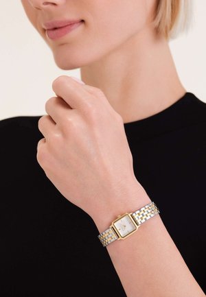 Montre-bracelet en métal argenté et doré avec un cadran carré blanc, un bracelet à maillons et des accents minimalistes, portée sur une main devant un fond sombre.