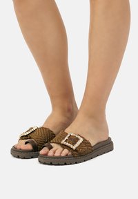 Las sandalias deslizantes trenzadas de color marrón presentan una gran hebilla decorativa con piedras de cristal transparente, una suela texturizada y una plantilla plana para mayor comodidad.