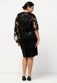 Capa de encaje negro con bordado floral, mangas semitransparentes y dobladillo curvado, llevada sobre un vestido negro de longitud a la rodilla. Tacones altos negros visibles por debajo.