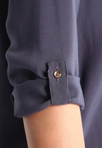 Dark gray fabric with a smooth texture, featuring a rolled cuff secured by a gold button. Close-up detail of sleeve construction.