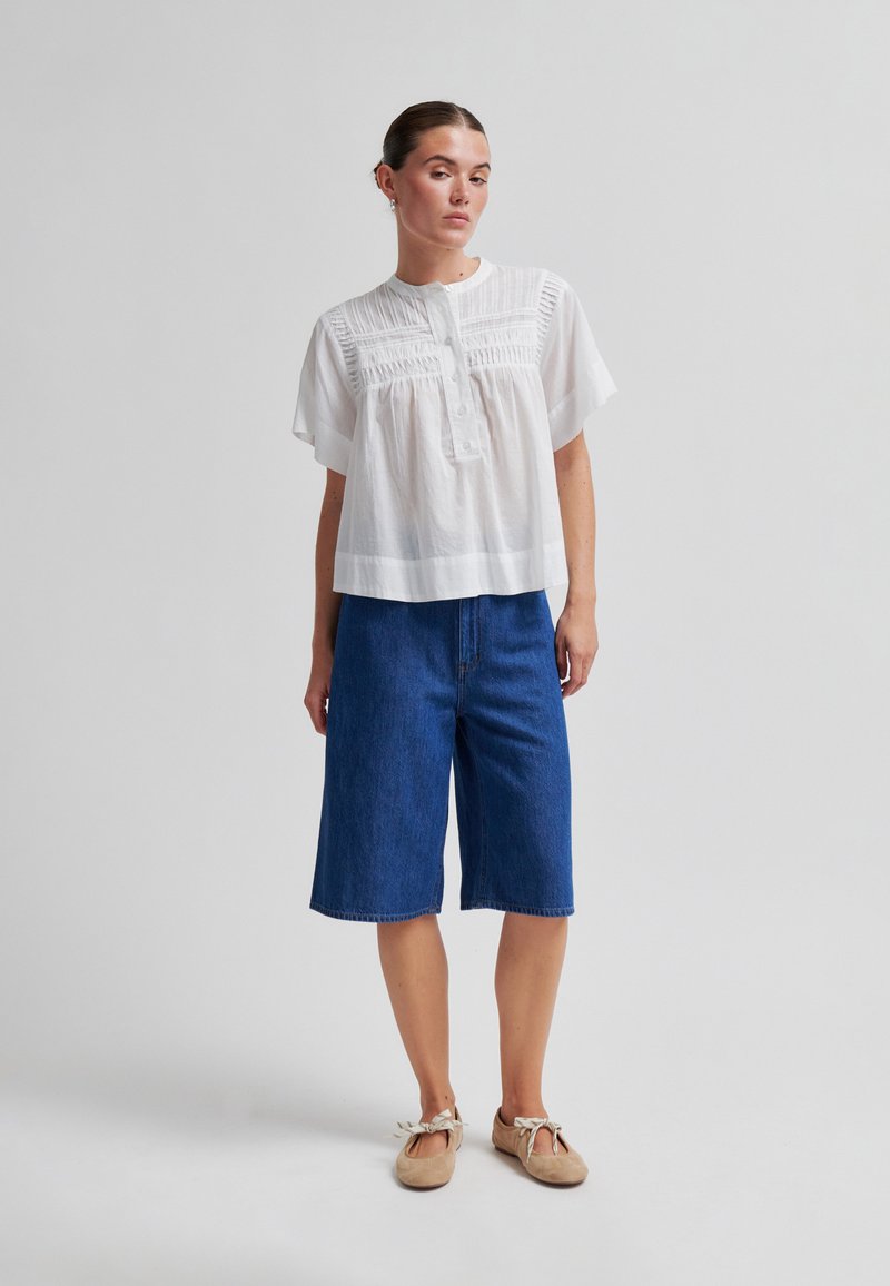 Femme portant un chemisier blanc à manches courtes avec des détails texturés, un short en denim bleu jusqu'aux genoux et des chaussures plates beige avec des liens autour de la cheville.