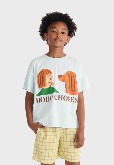 Dziecko o kręconych włosach nosi jasnoniebieską koszulkę z postacią kreskówkową dziewczynki i psa oraz żółte szorty w kratę, stoi z jedną ręką w kieszeni.