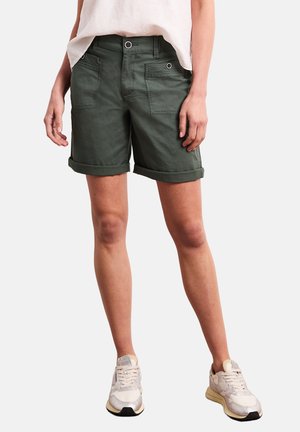 Persoon draagt opgerolde donkergroene shorts met voorzakken, een wit topje en witte sneakers, staand tegen een effen achtergrond.