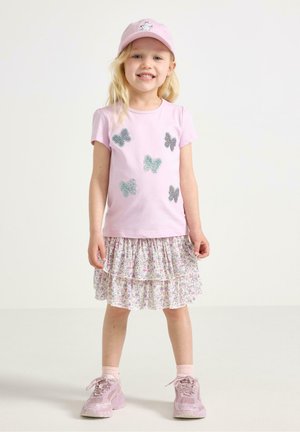 Giovane ragazza sorridente, indossa una maglietta rosa con una farfalla, gonna a più strati a fiori, cappellino rosa e sneakers rosa, in piedi davanti a uno sfondo semplice.