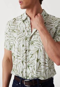 Camisa de manga corta con tela blanca y patrones de hojas verdes, cierre de botones y un ajuste relajado. Se lleva con un cinturón oscuro y vaqueros.
