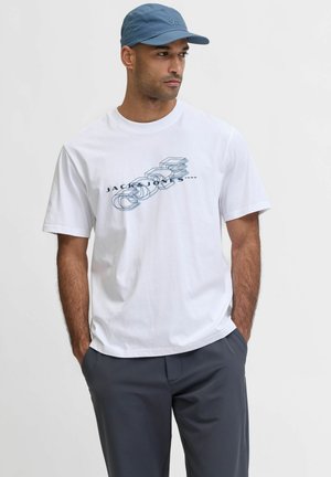 Biała bawełniana koszulka z nadrukiem w niebieskim kolorze z napisem "JACK & JONES 1990", w zestawieniu z szarymi spodniami i niebieską czapką.