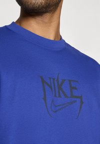 Mėlynas medvilninės medžiagos trumpomis rankovėmis marškinėliai su juodu Nike logotipo dizainu ir stilizuotu užrašu priekyje. Glotni tekstūra, laisvas kirpimas.