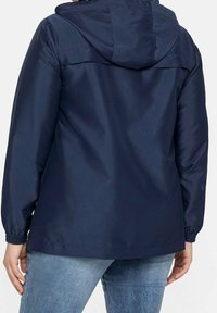 Giacca impermeabile blu navy con cappuccio, polsini elasticizzati e vestibilità comoda. Tessuto liscio e opaco con accenti minimi; adatta per un abbigliamento casual.