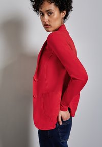 Röd blazer med skräddarsydd design, försedd med två knappar, en ficka och en slät, lätt glansig yta. Bärs med mörkblå jeans.
