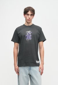 Młody mężczyzna w czarnej koszulce z fioletowym grafiką "Mniejszy Kawałek Przed Świętami" i jasnoniebieskich dżinsach, stojący na białym tle.