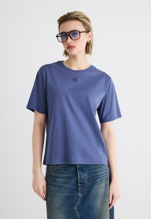 Vrouw met een blauw T-shirt met een klein logo, een denim rok en grote vierkante zonnebril, staand tegen een effen witte achtergrond.