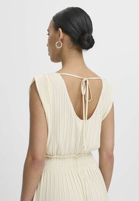 Kermakoski pleated dress, jossa on lyhyet hihat ja syvä V-mallinen selkäosa. Mekossa on solmittava kiinnitys ja kootut vyöt, jotka luovat ilmavan siluetin.