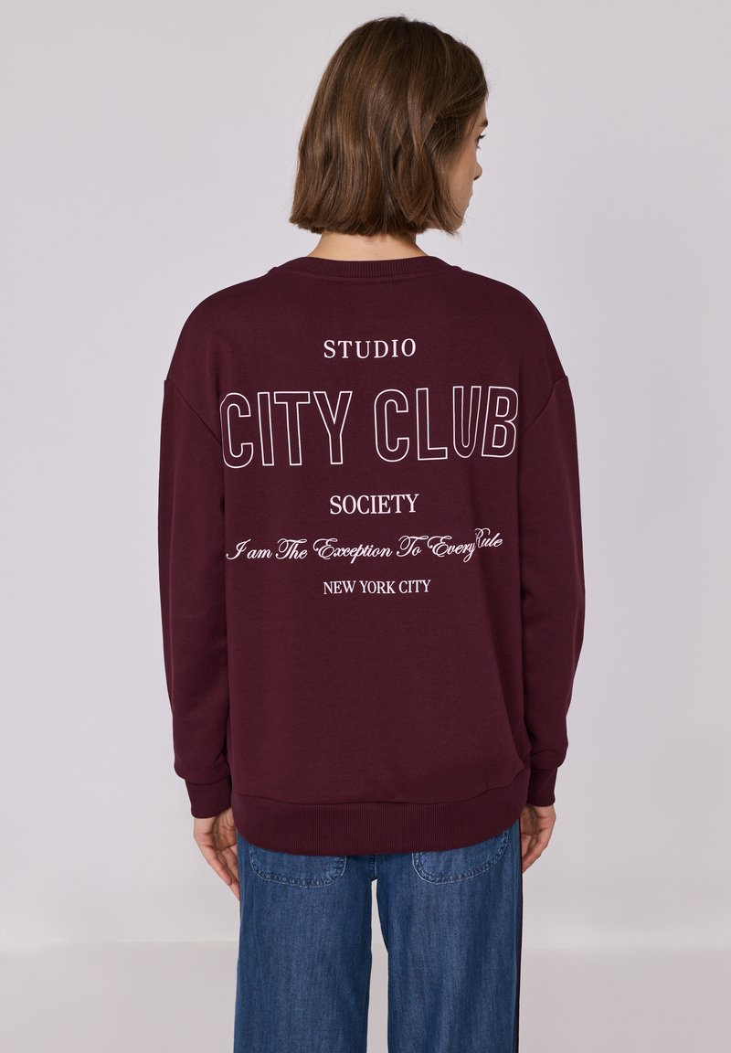 Sweatshirt bordeaux avec du texte blanc au dos disant "CITY CLUB" et "NEW YORK CITY", présentant une coupe décontractée et des poignets côtelés.