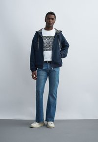 Mann stehend, trägt eine marineblaue Kapuzenjacke mit Reißverschluss, weißes T-Shirt mit Print, blaue Jeans, schwarzen Gürtel und weiße Sneaker vor schlichtem grauem Hintergrund.