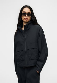 Γυναίκα με μακριά σκουρόχρωμα μαλλιά, φοράει μαύρο φαρδύ μπουφάν, μαύρο παντελόνι και ορθογώνια μαύρα γυαλιά ηλίου, στέκεται μπροστά σε μια απλή φόντο.