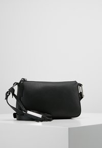 Black leather crossbody bag with adjustable strap and silver buckle, resting on a white surface against a gray background.