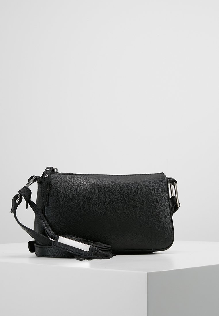 Black leather crossbody bag with adjustable strap and silver buckle, resting on a white surface against a gray background.