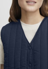 Marinblå kviltad väst med V-ringad design, som har texturerad sömnad och två framträdande tryckknappar, lagd över en ljusbeige topp.