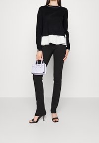 Czarny sweter z długim rękawem nałożony na biały top z falbanką, w połączeniu z czarnymi wąskimi spodniami i czarnymi szpilkami na pasku. Trzyma małą fioletową torebkę.