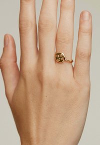 Anillo de oro con una cara redonda, que cuenta con cuatro acentos en forma de diamante. La banda tiene un diseño intrincado. Lleva en una mano con un tono de piel neutro.
