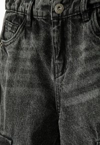 Vaqueros de mezclilla negra con un acabado texturizado, que cuentan con un botón plateado y un diseño clásico de cinco bolsillos. Detalle de costura visible a lo largo de las costuras.