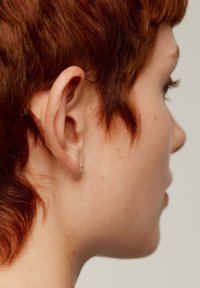 Pendiente en forma de estrella dorada exhibido en el lóbulo de la oreja, con un acabado pulido. Cabello corto y texturizado de color rojo visible de perfil.