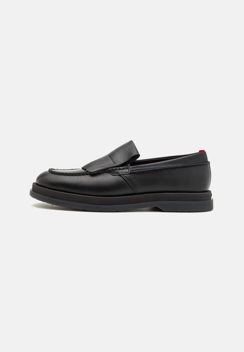 Mocassins noirs en cuir sans lacets avec des détails cousus et une semelle épaisse en caoutchouc, affichés sur un fond blanc, vue de côté.