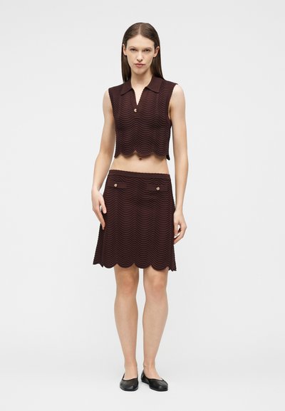 Femme portant un haut court sans manches en tricot marron et une jupe assortie mi-genoux avec un motif ondulé, ainsi que des ballerines noires, debout devant un fond blanc.