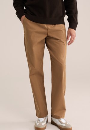 Homme portant un pantalon droit marron clair, un pull noir et des baskets blanches avec des accents beige, debout les mains détendues.