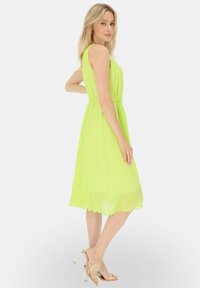 Bez rękawów, sukienka do kolan w jasnym neonowym żółtym kolorze z szyfonu, z dopasowanym wcięciem w talii, falbaniastym dołem i zestawiona z przezroczystymi obcasami.