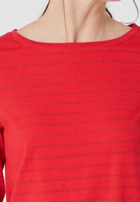 Maglietta rossa con strisce orizzontali, scollatura ampia e una texture morbida. Il tessuto ha una leggera lucentezza e una vestibilità comoda.