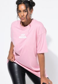 Kobieta z warkoczem nosi różowy, oversize'owy T-shirt z napisem "ICED MATCHA" oraz czarne skórzane spodnie, pozując na białym tle.
